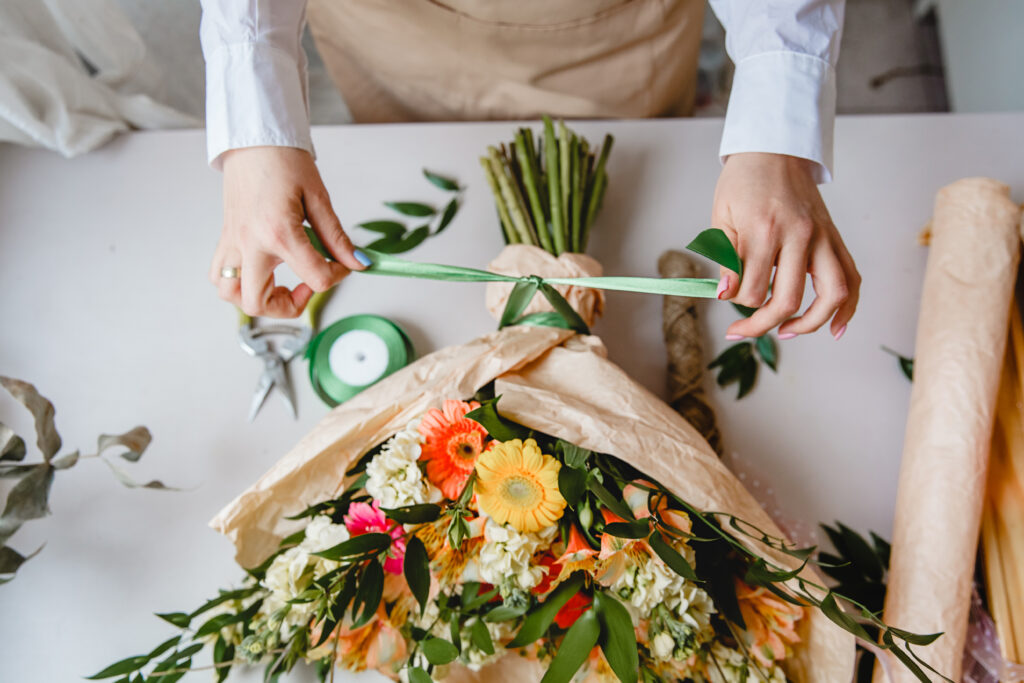 a female florist ties a ribbon bow on a bouquet of flowers wrapped in craft paper on the desktop. Top view. High quality photo